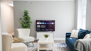 Ett modernt vardagsrum med en platt-TV som visar streamingtjänster, en vit soffa, en blå soffa, ett vitt soffbord och en stor krukväxt.