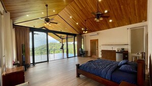 Quarto espaçoso com uma cama grande, teto de madeira e vista para uma paisagem exuberante através de amplas portas deslizantes.