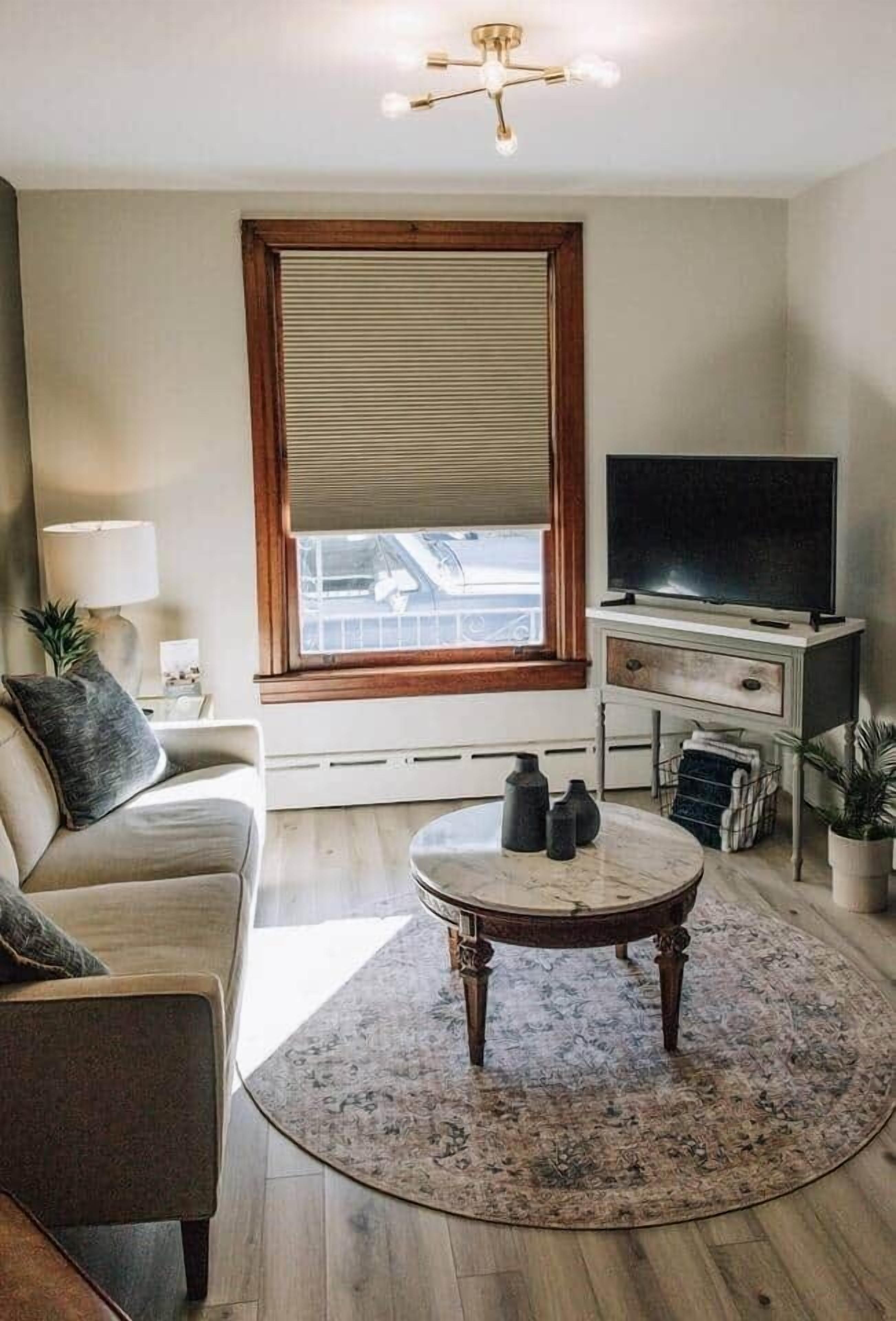 Ein Wohnzimmer mit einer Couch, einem runden Couchtisch, einem Fernseher auf einem Lowboard, einem Fenster mit Jalousien und einer Deckenleuchte.