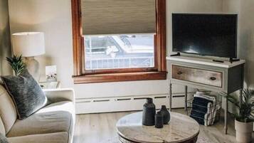 Ein Wohnzimmer mit einer Couch, einem runden Couchtisch, einem Fernseher auf einem Lowboard, einem Fenster mit Jalousien und einer Deckenleuchte.