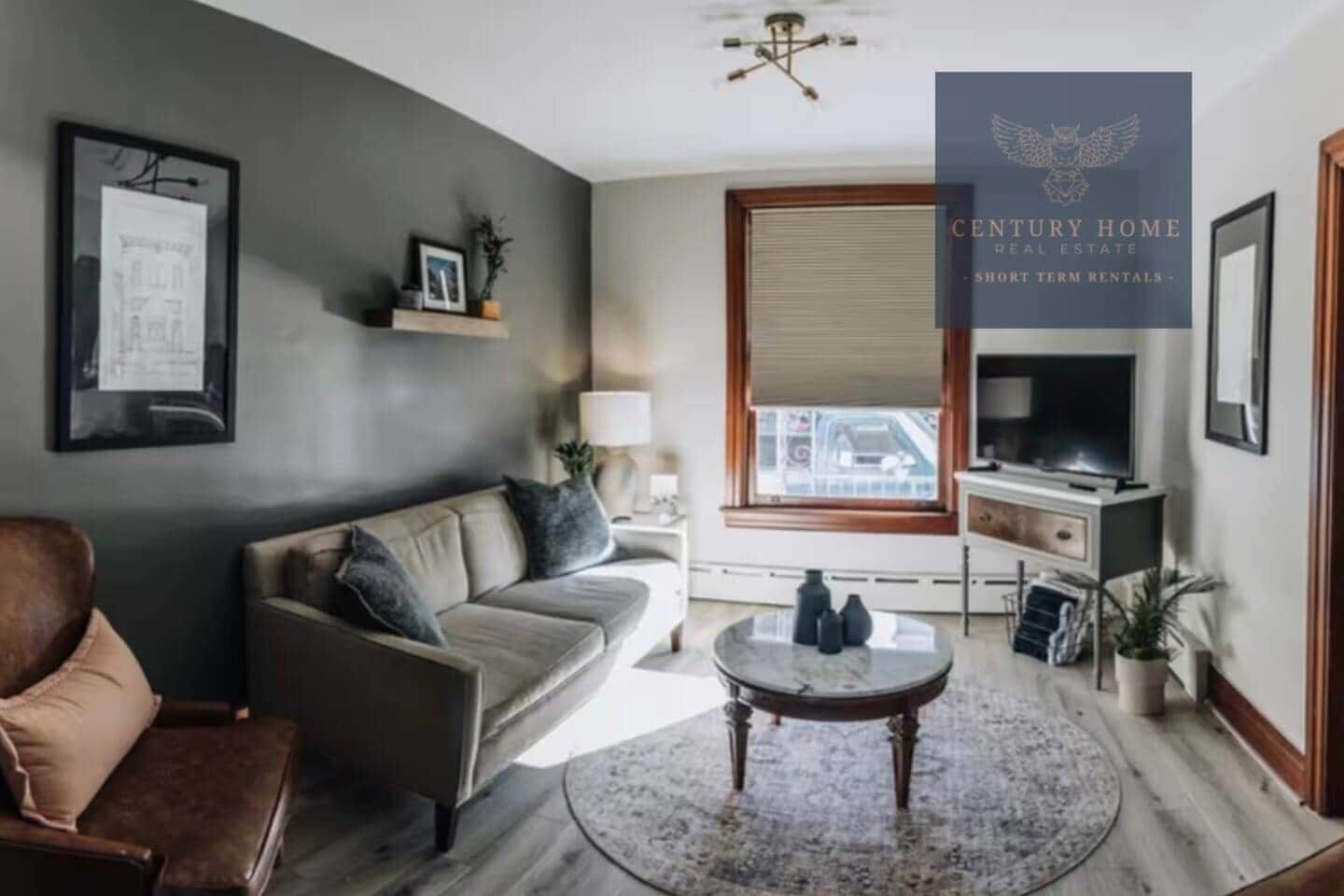 Un salon moderne avec un canapé, une table basse et un téléviseur. Il y a une fenêtre avec des stores, une lampe et un tableau encadré au mur.