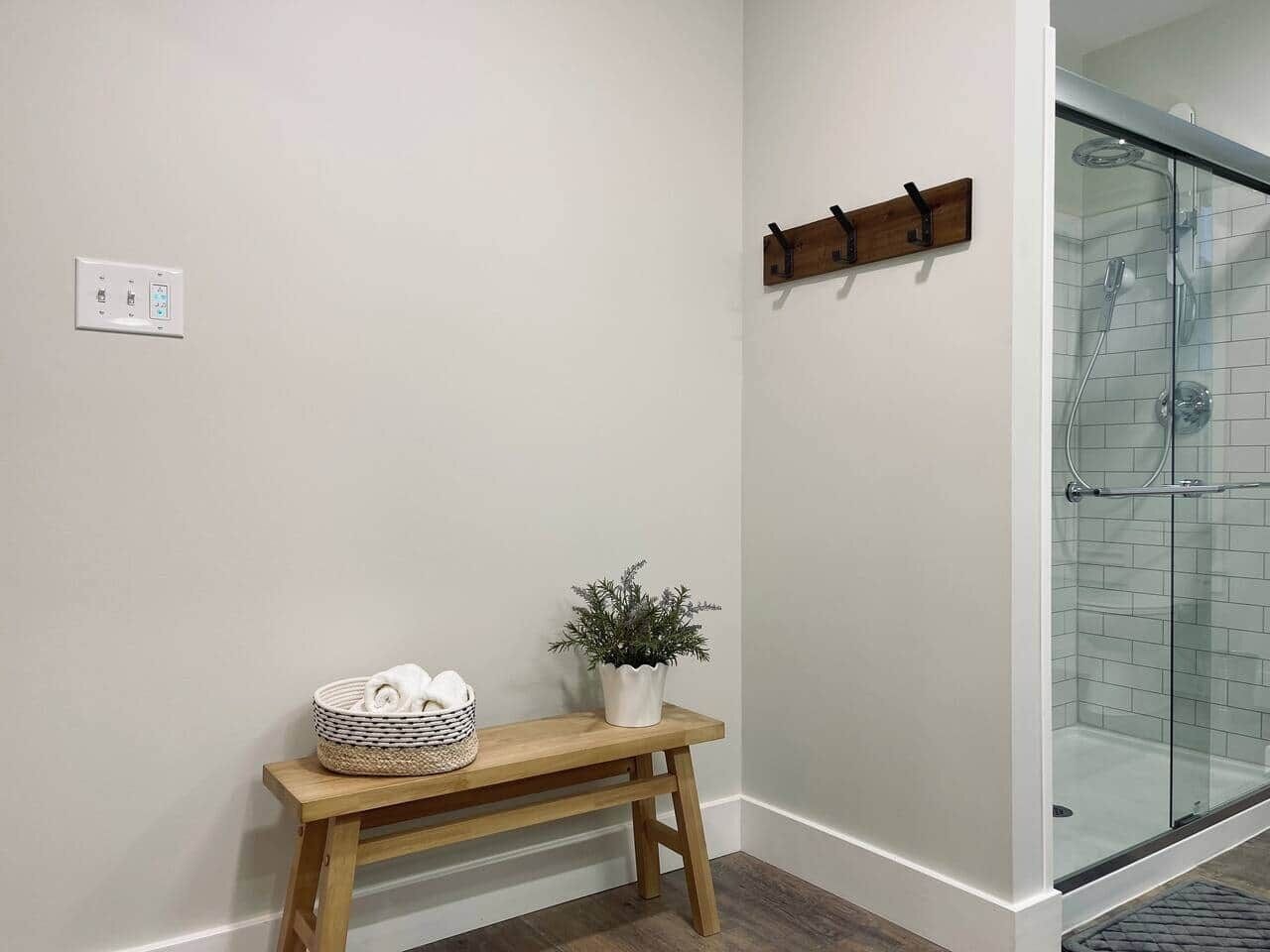 A bathroom with a glass shower enclosure, a wooden bench with a basket and plant, and a wall-mounted towel rack.