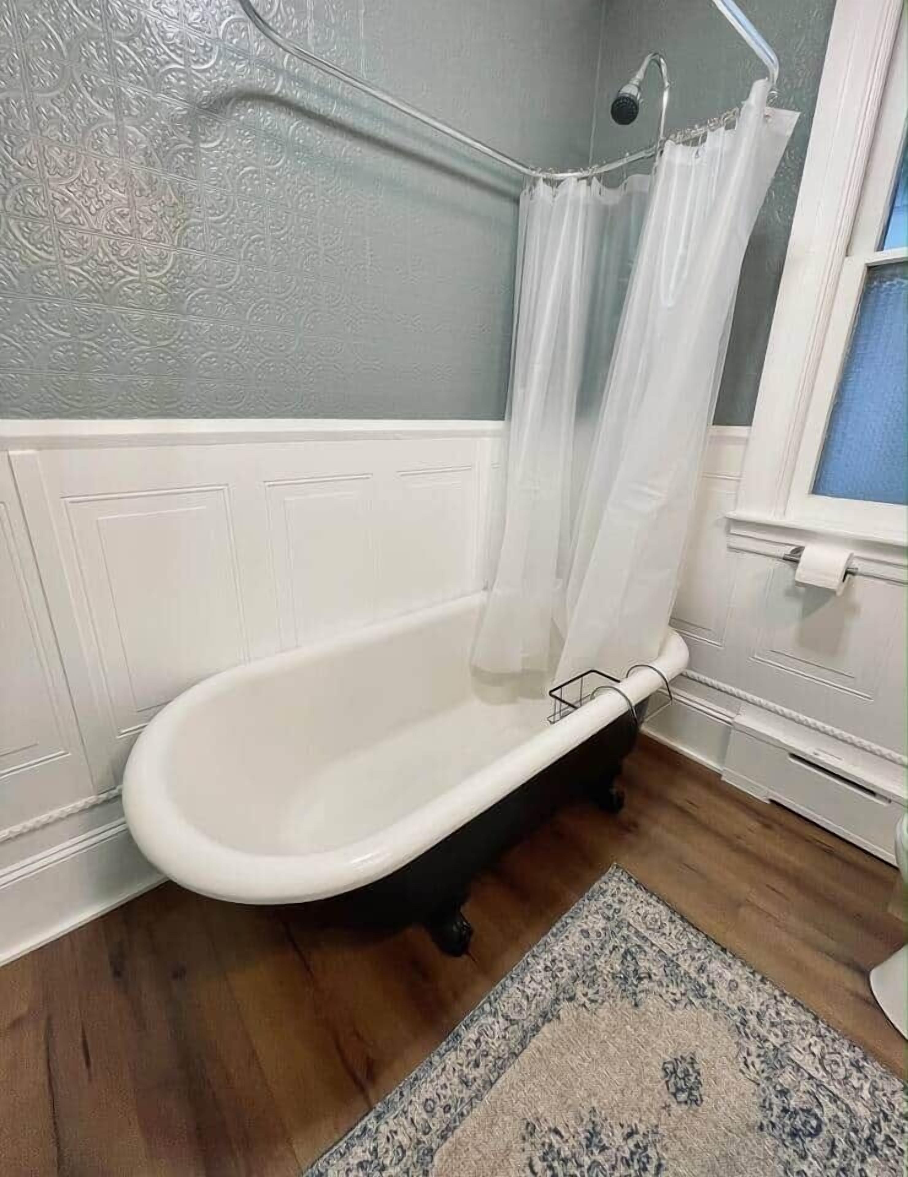 Een badkamer met een witte badkuip, een douchegordijn, een houten vloer en een vloerkleed met een patroon.