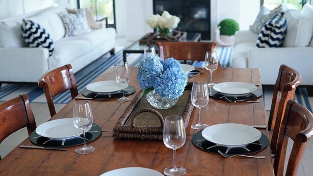 A dining area with a wooden table set for six, surrounded by wooden chairs. A vase with blue flowers is placed in the center.