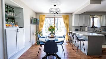 A modern kitchen with a dining area, featuring a central island, bar stools, and a view of the outdoors.
