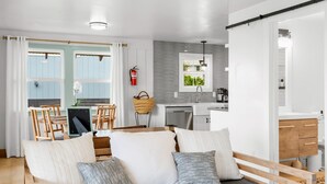 Uma sala de estar aconchegante com um sofá, almofadas e uma mesa de centro de madeira. Uma cozinha é visível ao fundo, com uma janela e um extintor de incêndio.
