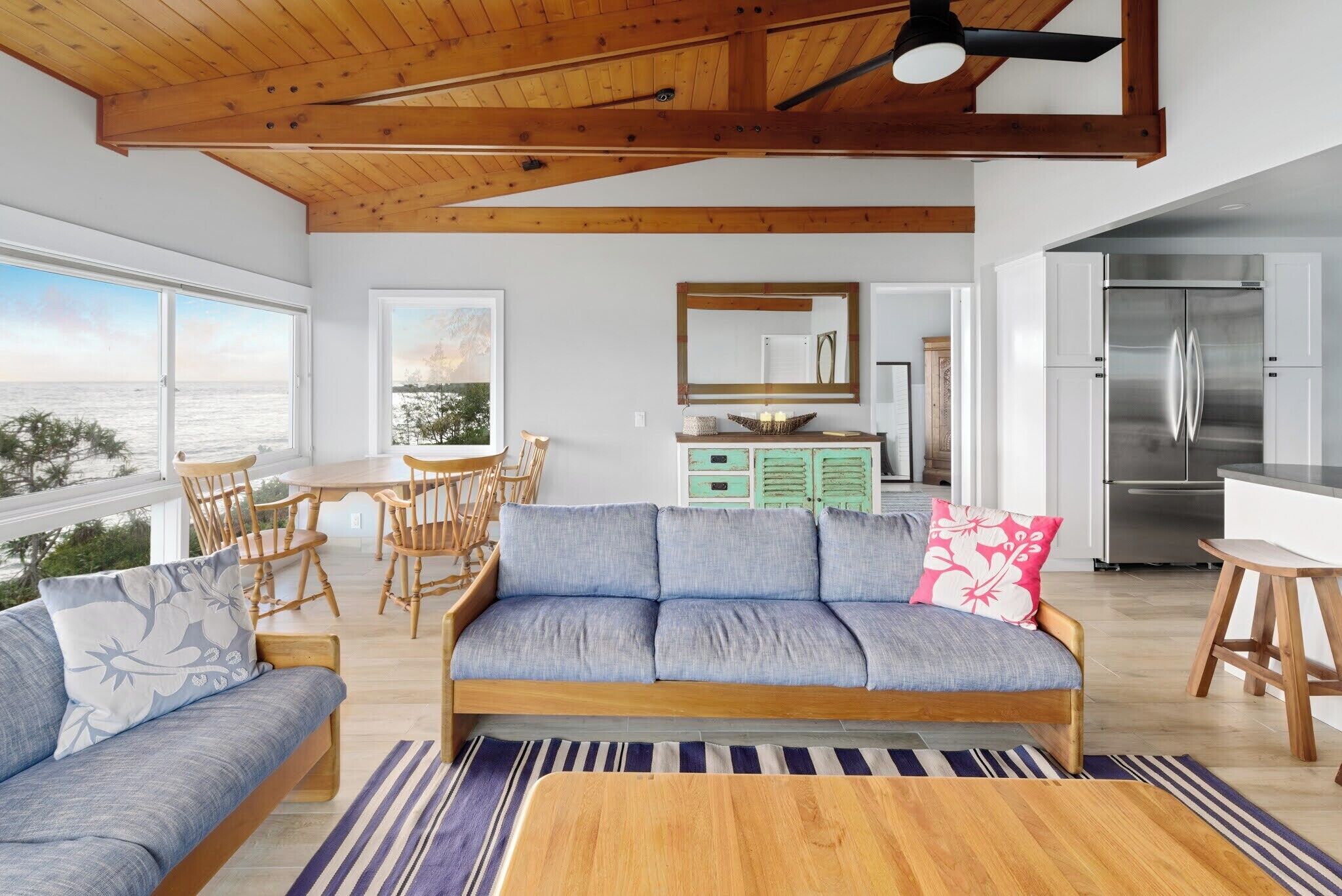Una sala de estar moderna con sofá, mesa de comedor y sillas, una cocina con refrigerador, y vistas al mar.