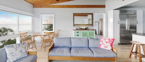 Una sala de estar moderna con sofá, mesa de comedor y sillas, una cocina con refrigerador, y vistas al mar.