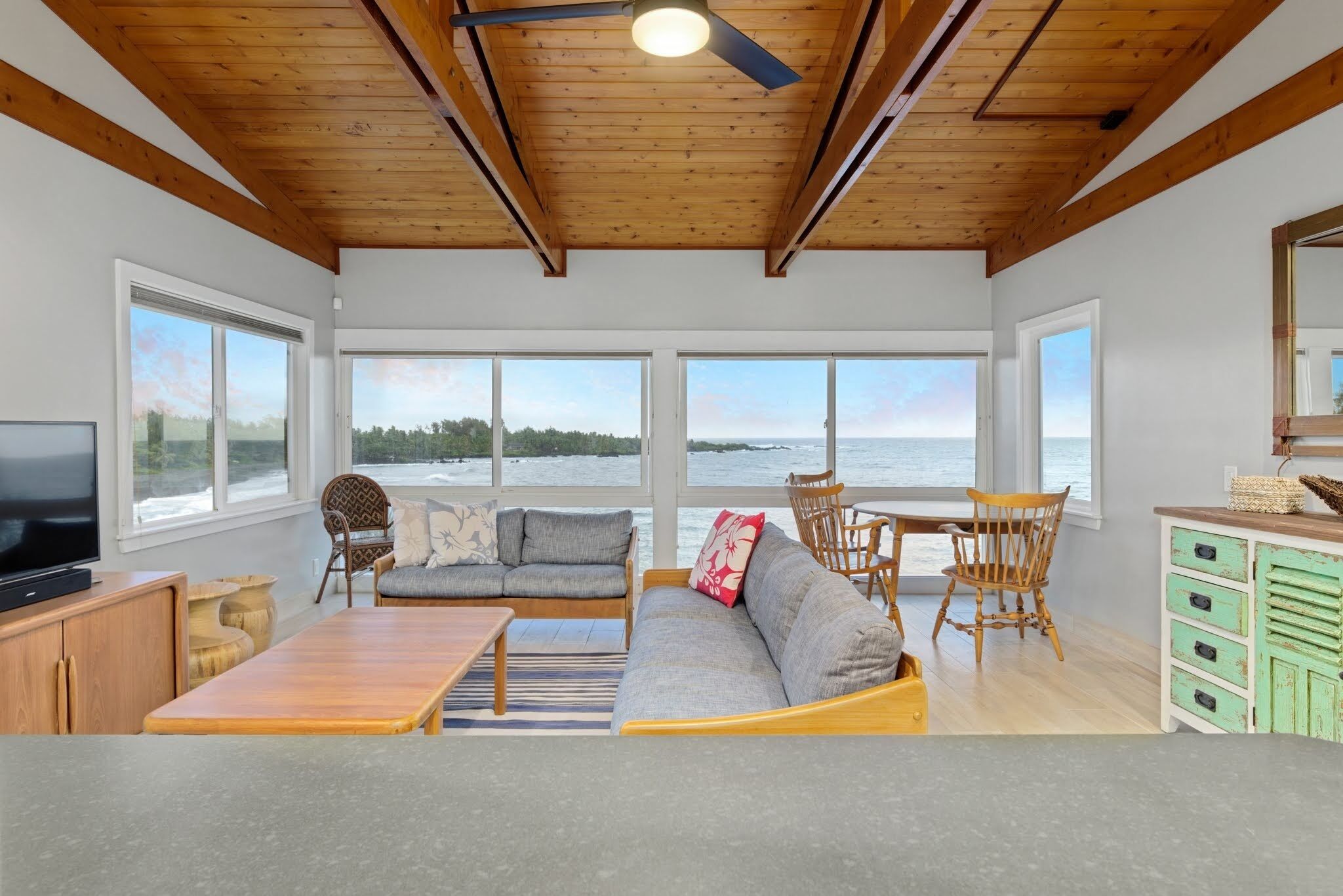 Amplia sala de estar con techo de madera, ventanas grandes con vista al mar, un sofá, sillas, una mesa de madera y un televisor.