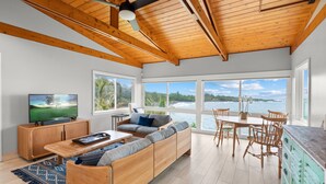 Una sala de estar moderna con techo de madera, un ventanal con vistas a una playa, un sofá, una mesa de centro y un televisor sobre un mueble de madera.