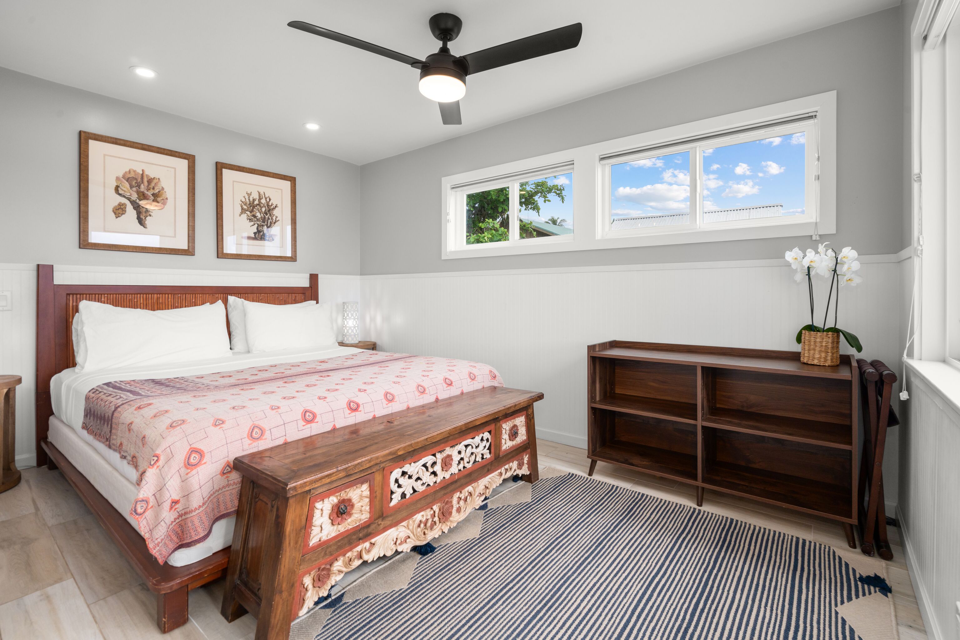 A bedroom with a bed, a wooden bench, a ceiling fan, and a window with a view of the sky.