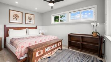 A bedroom with a bed, a wooden bench, a ceiling fan, and a window with a view of the sky.