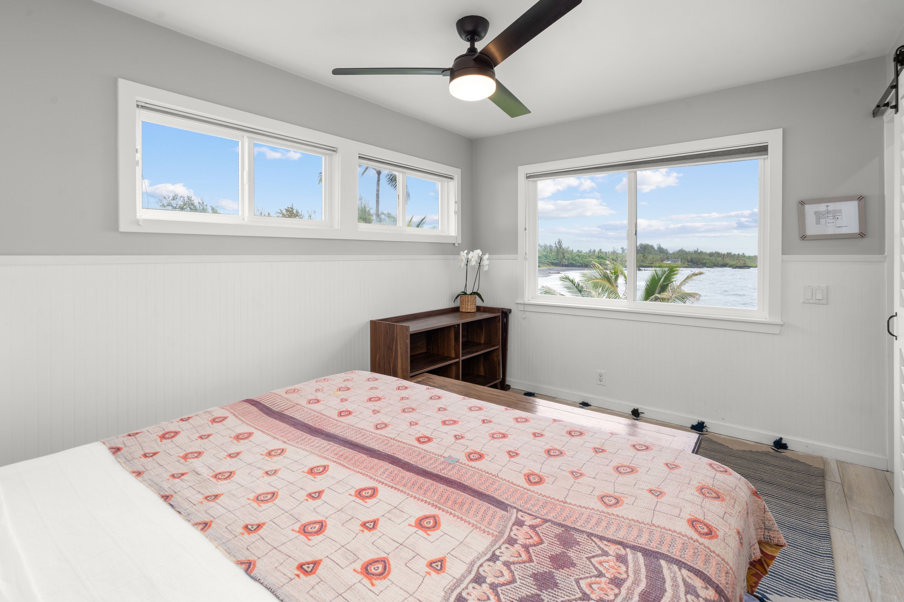 A bedroom with a bed, two windows, a ceiling fan, and a view of the water.