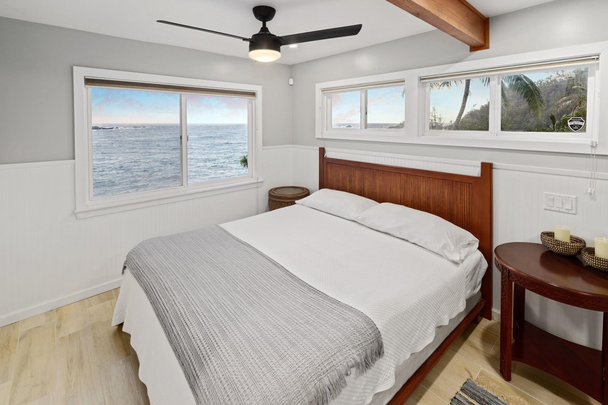 A bedroom with a bed, a ceiling fan, two windows with views of the sea, and a small wooden table with candles.