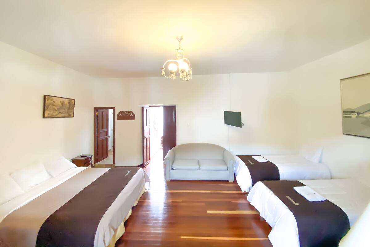 Ein Hotelzimmer mit drei Betten, Holzboden, einer Chandelüster und einem Gemälde an der Wand.