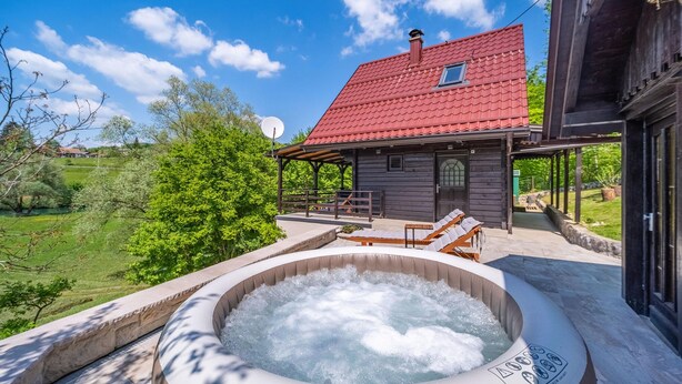 A hot tub with a wooden deck, a wooden cabin with a red roof, and a green landscape in the background.