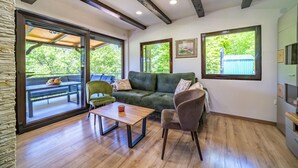 A modern living room with a green sofa, a wooden coffee table, and a grey armchair. There is a large window with a view of greenery outside.