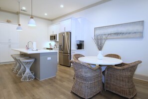 A modern kitchen with white cabinetry, stainless steel appliances, a central island with bar stools, and wicker chairs around a small table. - A Short walk to 30A beach -Pool/Bikes/Arcade games (Walton County)
