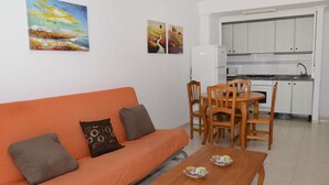 A living room with an orange sofa, wooden dining table, and chairs. A kitchen with white cabinets and appliances is visible in the background.
