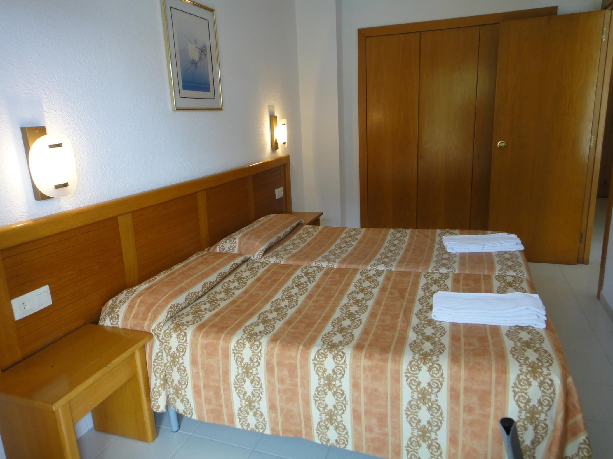 Uma cama bem arrumada com colcha listrada, uma mesa de cabeceira de madeira e um guarda-roupa com porta.