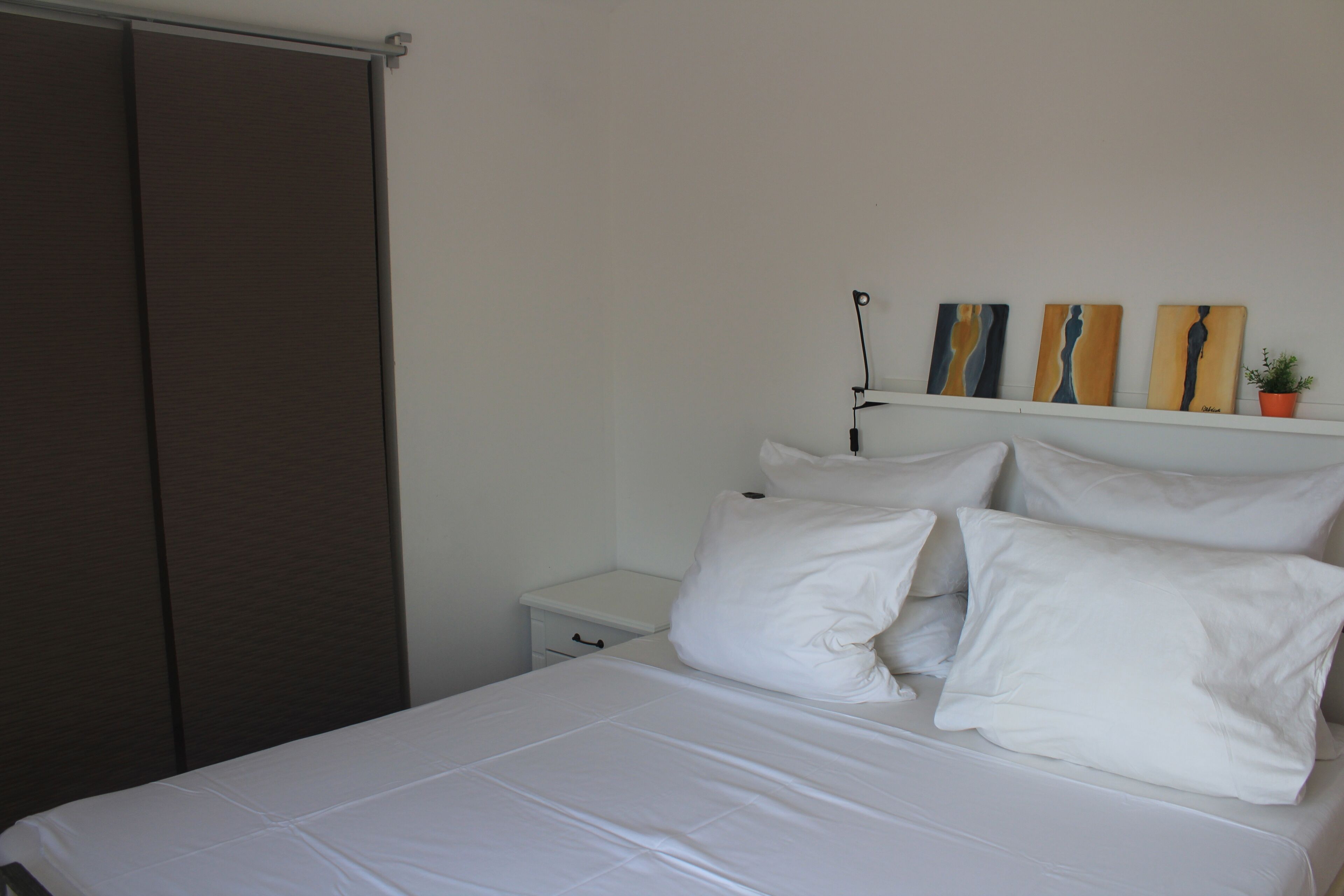 A neatly made bed with white linens and pillows, a white bedside table with a drawer, and a wall-mounted shelf with three artworks and a small potted plant.