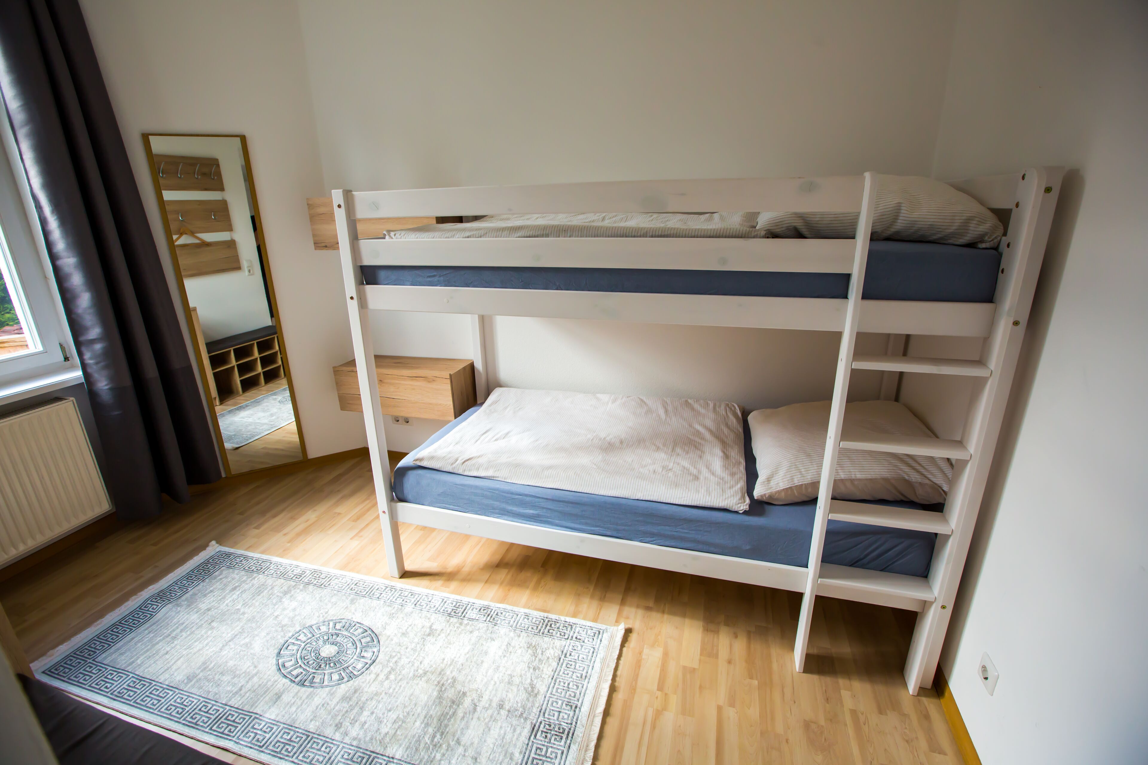 Un lit superposé avec un lit en bas et un lit en haut, un miroir, une commode en bois et un tapis à motifs.