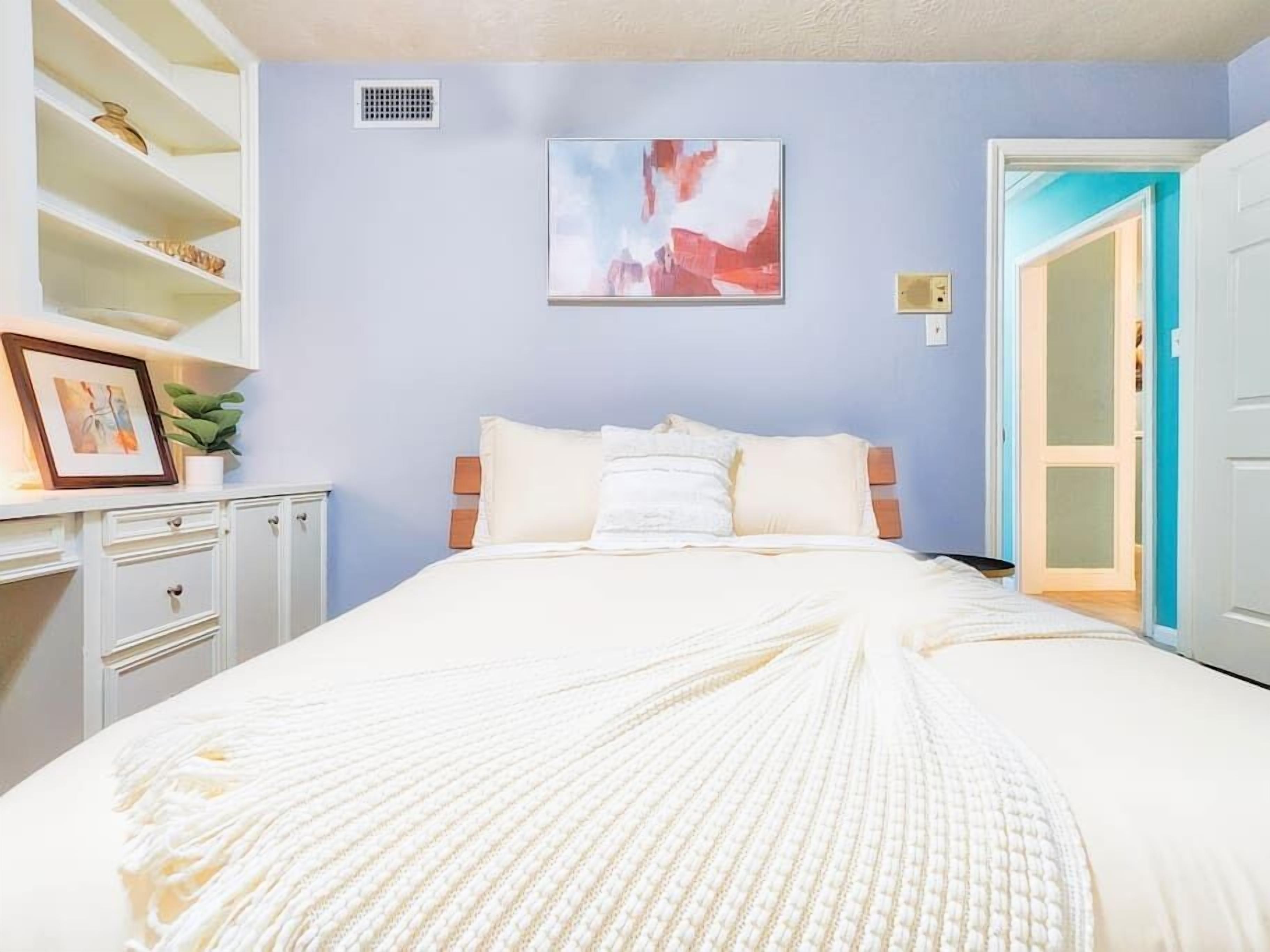 Quarto com cama, mesa de cabeceira, prateleira com objetos decorativos e porta que dá para outro cômodo.