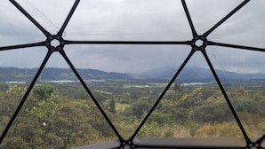 A view through a geometric window structure, revealing a landscape with trees and distant hills.