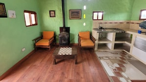 A cozy living room with a high ceiling, a fireplace, two wooden chairs with cushions, a wooden coffee table, and a kitchen area with a sink and stove.