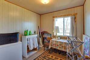 A room with wooden walls, a window with curtains, a table with a patterned tablecloth, a lamp, a drying rack, a washing machine, and a basket. - Charming 'Corral Creek Ranch House' in Circle (Circle)