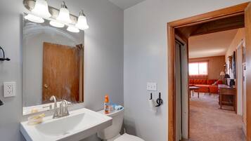 A bathroom with a white sink, mirror, and toilet. There is a shower area with lights and a wooden door. A hallway leads to a living room with a red sofa and a table.