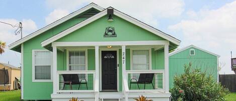 A green house with a porch, two chairs, and a small garden.
