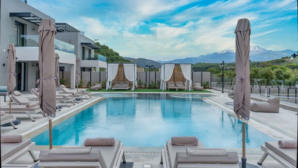 A modern pool area with lounge chairs and umbrellas, a clear sky, and a mountainous backdrop.