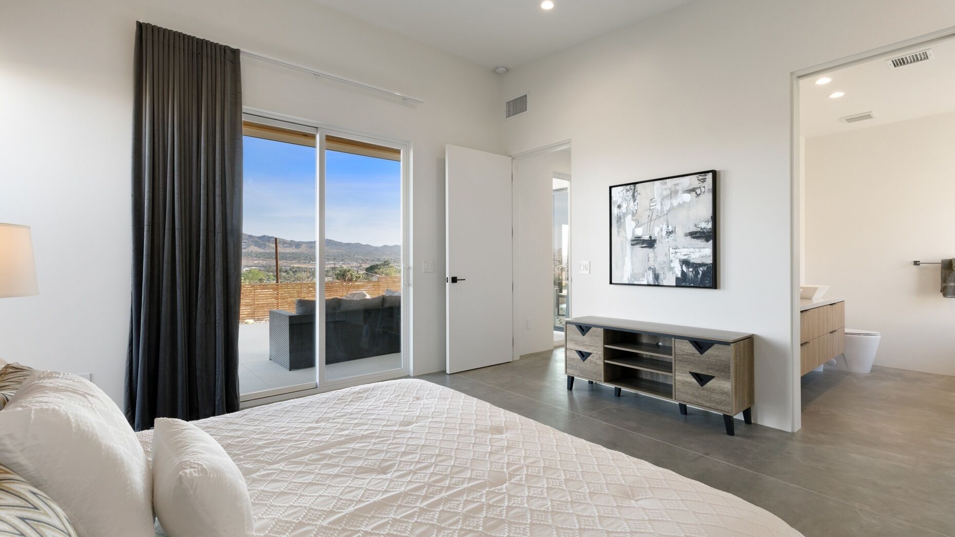 A bedroom with a large bed, a sliding glass door to a balcony, a modern dresser, and a framed abstract painting on the wall.