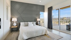 Un dormitorio moderno con una cama grande, dos mesitas de noche con lámparas, vistas a un paisaje desértico a través de amplios ventanales y un diseño geométrico en la pared.