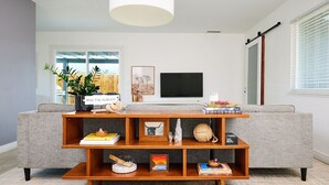 A modern living room with a grey sofa, a wooden coffee table with books and decorative items, a flat-screen TV, and a large window with blinds. - Live Centered W/ Hot Tub, Fire Pit In Joshua Tree 2 Bedroom Home by RedAwning (Joshua Tree)
