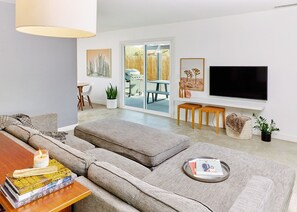 Un salon moderne avec un canapé d’angle gris, une table basse avec des livres, un téléviseur fixé au mur et une porte-fenêtre coulissante donnant sur une terrasse extérieure.