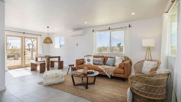 A spacious living room with a sofa, coffee table, and wicker chairs. A dining area with a table and chairs is visible. Large windows offer a view of the outdoors. - Mojave Mesa - Desert Views & Desert Style 1 Bedroom Home by RedAwning (Yucca Valley)