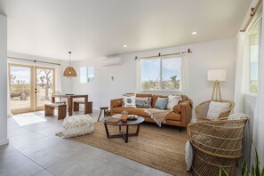 Un salón amplio con sofá, mesa de centro y sillas de mimbre. Se aprecia un comedor con mesa y sillas. Grandes ventanales ofrecen vistas al exterior.