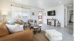 A modern living room with a leather sofa, a round coffee table, and a white pouf. There is a television mounted on the wall and a wicker chair. - Mojave Mesa - Desert Views & Desert Style 1 Bedroom Home by RedAwning (Yucca Valley)