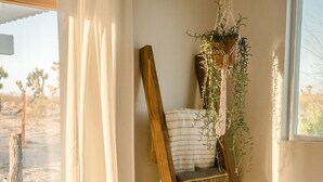 A room with a wooden chair, a hanging plant, a potted plant, and a view of a desert landscape through a window. - Mojave Mesa - Desert Views & Desert Style 1 Bedroom Home by RedAwning (Yucca Valley)