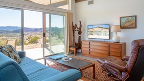 A modern living room with a blue sofa, a leather chair, a wooden coffee table, and a view of the desert landscape through large sliding glass doors. - Las Alas Del Sol- A Desert Architectural Gem 2 Bedroom Home by RedAwning (Joshua Tree)