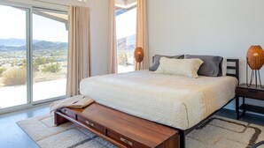 A bedroom with a large bed, a wooden nightstand, a woven lamp, and a view of a mountainous landscape through a sliding glass door. - Las Alas Del Sol- A Desert Architectural Gem 2 Bedroom Home by RedAwning (Joshua Tree)