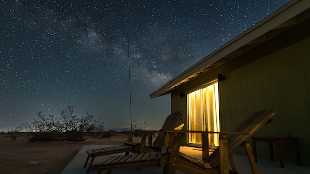 Un ciel étoilé offrant une vue sur la Voie lactée, une terrasse en bois avec des chaises longues et un bâtiment dont l’intérieur est éclairé.
