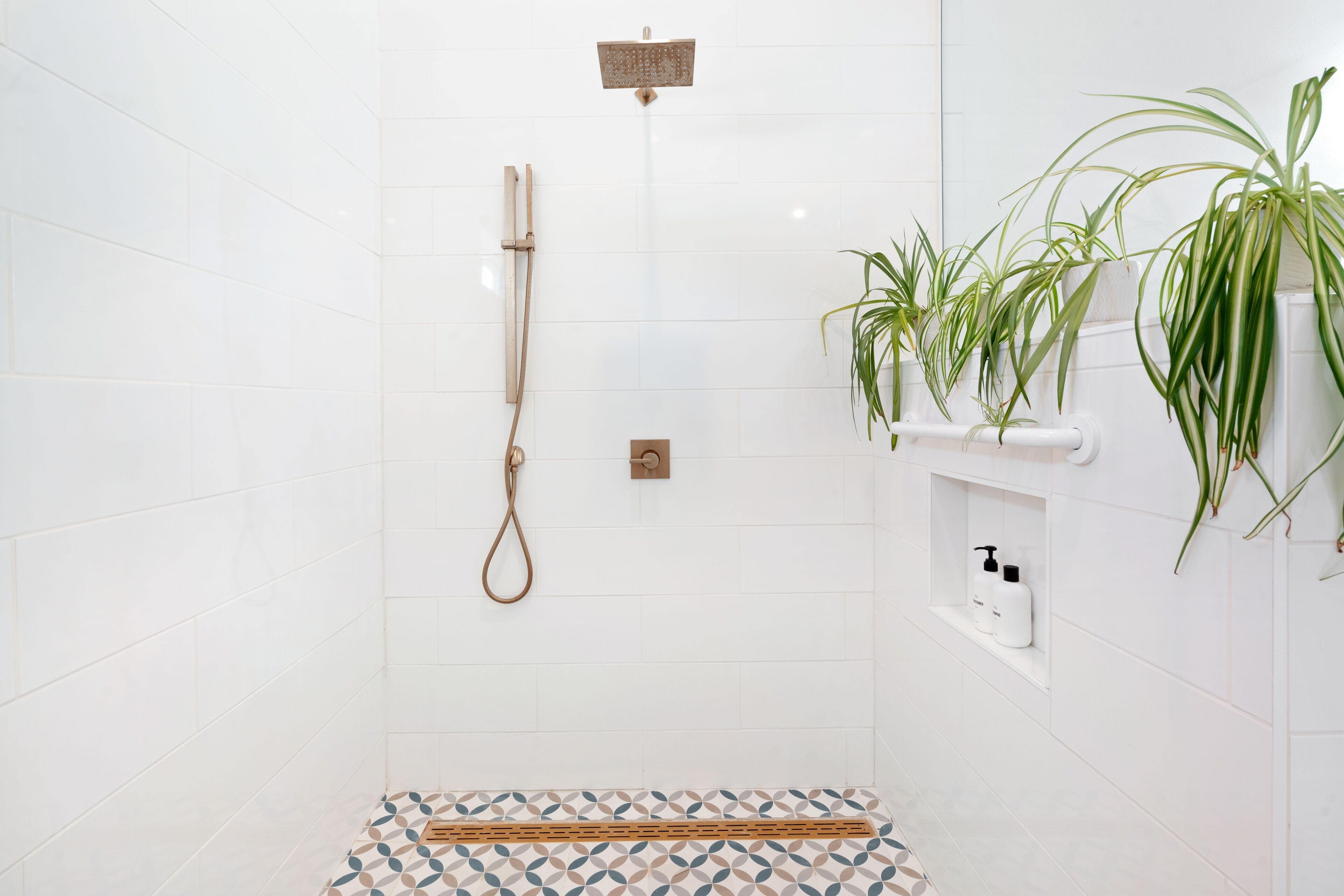 A modern bathroom with white tiles, a shower with a handheld showerhead, a wooden bath mat, and a shelf with toiletries.