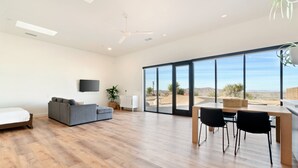A spacious living area with a wooden floor, a gray sofa, a dining table with chairs, and a large window offering a view of the desert. - Back Haus - Views, Dark Skies And Fire Pit! 1 Bedroom Home by RedAwning (Joshua Tree)