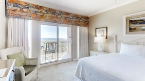 A hotel room with a large bed, a chair, a nightstand, a lamp, and a view of the ocean through a sliding glass door. - Tops'l 1008a Chalet by RedAwning (Miramar Beach)