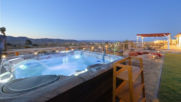 A hot tub with a wooden deck and a pergola with string lights, overlooking a cityscape at dusk. - Rancho Nopales Jt - Pool, Hot Tub, Hammocks, Fast Wifi + Views! 4 Bedroom Home by RedAwning (Joshua Tree)