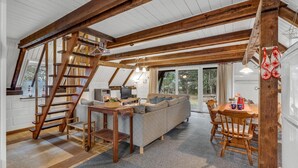 Una sala de estar estilo loft acogedora, con cama en entrepiso, vigas de madera, sofá, mesa de comedor y televisor.