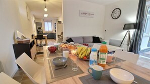A modern living room with a dining area, featuring a sofa, a coffee table with fruits and drinks, and a kitchen in the background. - Trémélois - TREMEL - Gîte (TREMEL)